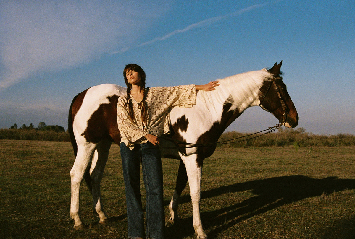 Woman in a lace top resting her hand on a paint horse in a field